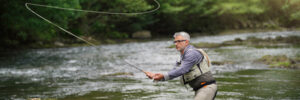 Man standing in a river fly fishing, surrounded by green trees and sunlight filtering through the foliage.