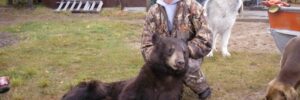 A woman in camouflage kneels behind a dead bear outdoors, with two dogs nearby and camping equipment in the background.