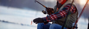 Older man in winter clothes fishing from a boat on a calm, cold lake, holding a rod with a lure.