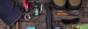 Fishing gear, including rods, reels, lures, boots, a hat, pliers, knife, GPS, and tackle box, arranged on wood.