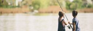 Two young children stand by a lake, fishing together with a single fishing rod on a grassy shoreline.
