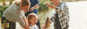 Two adults and two children by a lake, with one child holding a fish and the others watching and smiling.
