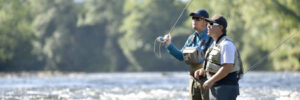 Two people wearing waders and hats are fly fishing while standing in a shallow, flowing river surrounded by trees.