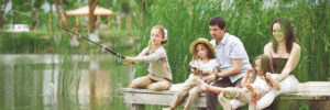 Family of five sitting on a wooden dock fishing, surrounded by greenery and a calm lake.