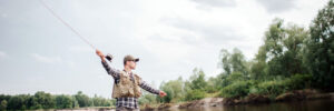 Person standing in shallow river water fly fishing, surrounded by trees under a cloudy sky.