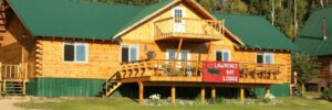 Wooden lodge with green roof, front porch, and a red banner reading "Lawrence Bay Lodge," set among trees and grass.