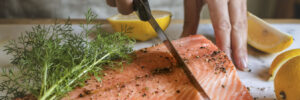 Person slicing a seasoned raw salmon fillet with a knife on a cutting board, with dill and lemon wedges nearby.