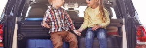 Two children sit on luggage in the open trunk of a car, holding hands and looking at each other.