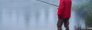 Person in a red raincoat fishing by a misty lake, surrounded by trees and grass.