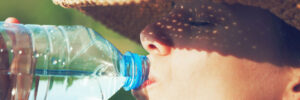 Person wearing a straw hat drinks water from a plastic bottle outdoors on a sunny day.
