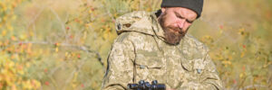 A man in camouflage clothing outdoors holds and checks a rifle with a scope, surrounded by foliage.