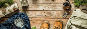 Camping gear arranged on a wooden surface, including boots, jacket, backpack, camera, thermos, mug, and accessories.