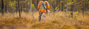 A person in orange camouflage gear stands in a forest clearing holding a rifle, surrounded by trees and grass.