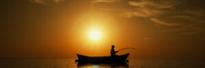 Silhouette of a person fishing in a small boat on calm water at sunset, with the sun low on the horizon.