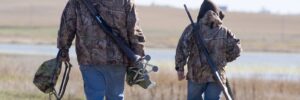 Two people in camouflage jackets walk through a field carrying rifles and gear, with open land and water in the background.