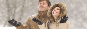 Two people in winter coats stand outside smiling and looking up as snow falls around them.