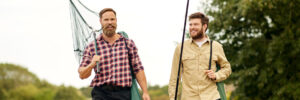Two men walk outdoors carrying fishing gear, including a net, a bucket, and a fishing rod, with trees in the background.