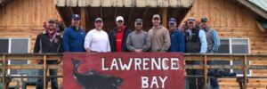 Ten men stand on a wooden balcony behind a large "Lawrence Bay Lodge" sign with a fish logo.