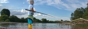 A child wearing a hat and yellow boots stands in shallow water while fishing with a rod in a river.
