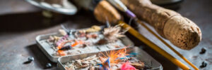 Close-up of fishing rods, reels, and an open box of assorted fishing flies on a table.