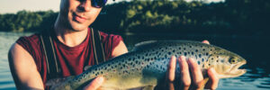Person wearing sunglasses and a cap holds a freshly caught fish on a boat with water and trees in the background.