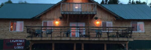 A wooden lodge with a balcony and a sign reading "Lawrence Bay Lodge" is lit up at dusk, surrounded by trees.