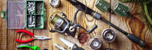 Fishing tackle and equipment, including a rod, reels, pliers, knife, scissors, weights, and hooks, arranged on a wooden surface.