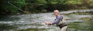 Man standing in a shallow river, casting a fishing line with a fly fishing rod, surrounded by green trees.