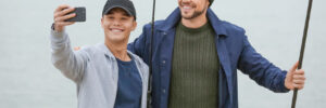 Two people holding fishing rods take a selfie together by the water on a cloudy day.