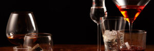 Assorted glasses of alcoholic beverages, some with ice, arranged on a wooden surface against a black background.