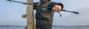 Person holding a large fish and fishing rod while standing on a boat in open water on a clear day.
