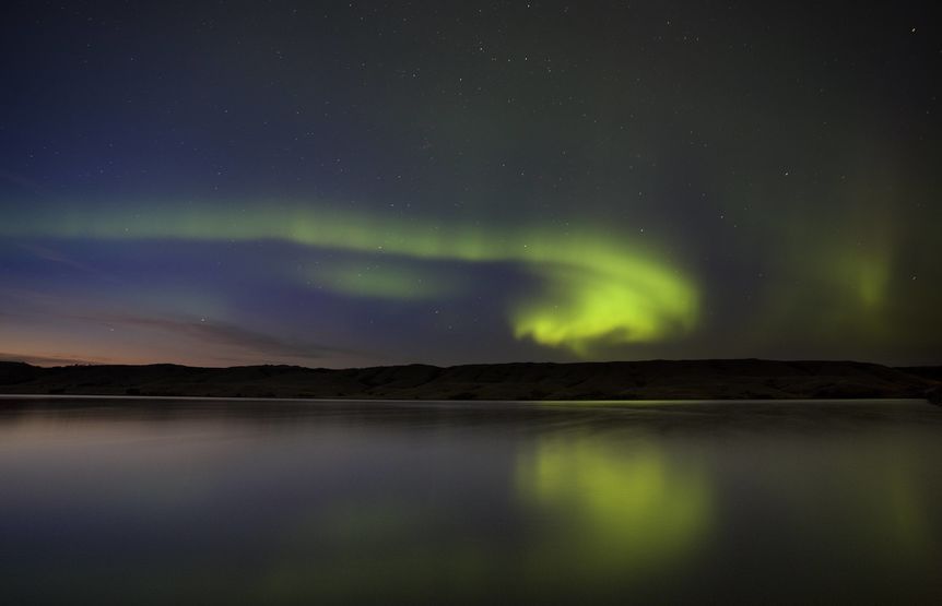 Green aurora borealis lights streak across the night sky, reflected in a calm body of water with distant hills.