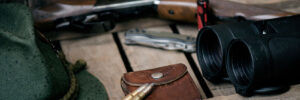 A hunting rifle, two bullets, green hat, binoculars, multitool, and leather pouch on a wooden surface.