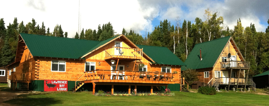 Two log cabins with green roofs sit on a grassy lawn near a forest; a sign reads "Lawrence Bay Lodge.