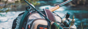 Fishing gear, including a backpack and fishing rod, resting on grass by a river in a wooded outdoor setting.