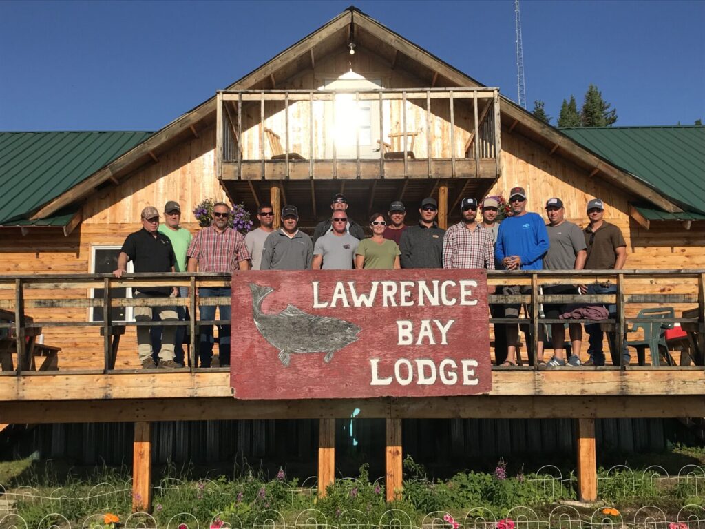 A group of people stand on the deck of Lawrence Bay Lodge in front of a large sign on a sunny day.