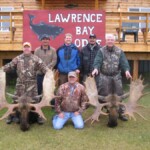 Six men posing with two large moose antlers at Lawrence Bay Lodge, with a log cabin in the background.