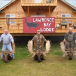 Three people standing with two large moose antlers in front of a wooden lodge labeled "Lawrence Bay Lodge.