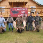 Six men hold moose antlers in front of a wooden lodge with a sign reading "Lawrence Bay Lodge" in the background.