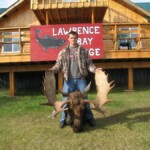 Person holding moose antlers in front of a lodge with a sign reading "Lawrence Bay Lodge.