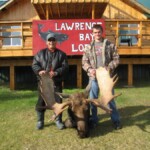 Two people standing behind a large moose they hunted, on the grass in front of a wooden lodge with a "Lawrence Bay Lodge" sign.