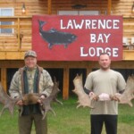 Two men hold large moose antlers in front of a sign for Lawrence Bay Lodge, standing by a wooden building.