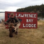 Person kneeling behind a moose head in front of a "Lawrence Bay Lodge" sign with a lake and trees in the background.