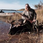 Person in camouflage holding a rifle, sitting on a moose by a lakeside with a boat in the background.