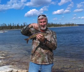 Man in camouflage jacket holding a fish by a lakeshore, with blue sky and clouds in the background.