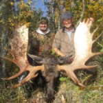 Two people pose with a large moose in a forested area, holding its antlers. Date on the image is September 21, 2010.