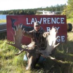 Person posing with large moose antlers in front of a "Lawrence Bay Lodge" sign, near a lake with a floatplane visible.
