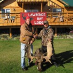 Two people shaking hands in front of Lawrence Bay Lodge, with a large moose antler displayed on the grass.