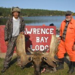 Two men stand with a harvested moose in front of a "Wrence Bay Lodge" sign by a lakeside with boats and trees in the background.