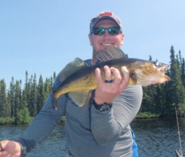 Person holding a fish while standing on a boat near a forested lake, wearing a cap and sunglasses, smiling at the camera.
